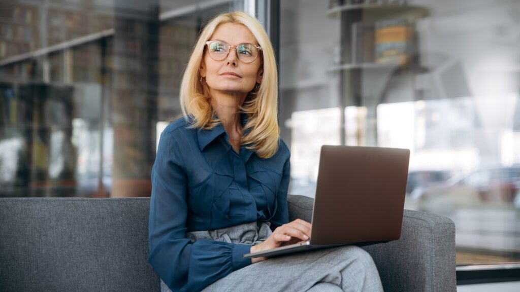 Mature caucasian confident business woman wearing eyeglasses and a stylish blue shirt sitting on a sofa with a laptop and looking away